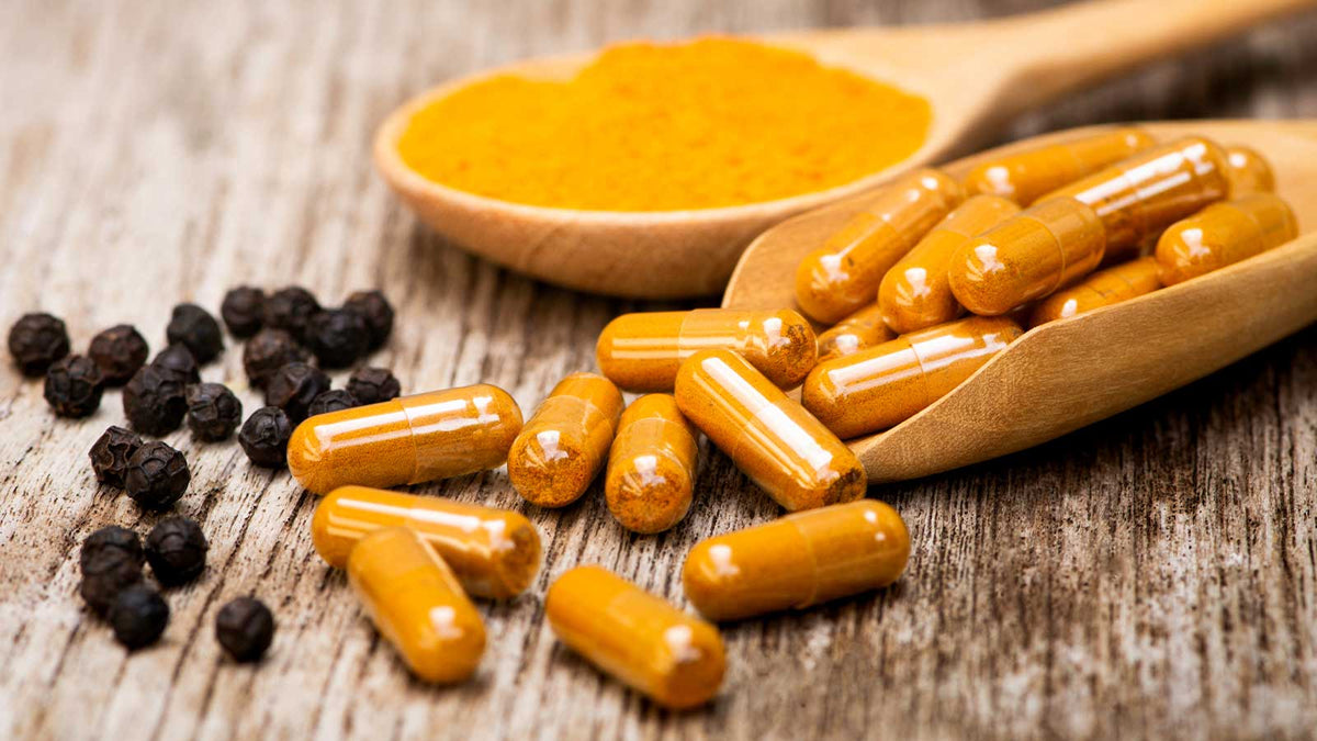 Curcuma-Kapseln und schwarze Pfefferkugeln (Piperin). Ein Holzlöffel gefüllt mit Curcuma im Hintergrund