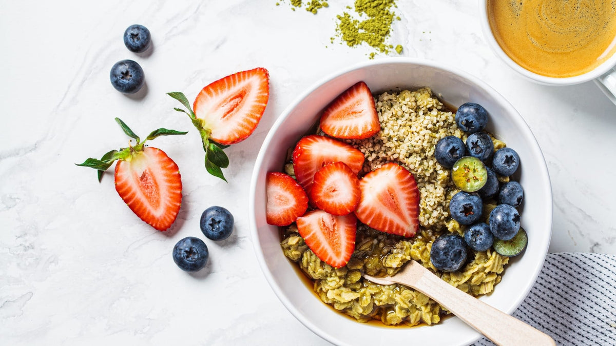 Frühstücksmüsli mit Erdbeeren und Blaubeeren sowie Hanfproteinpulver auf hellem Untergrund mit Holzlöffel neben Kaffeetasse.
