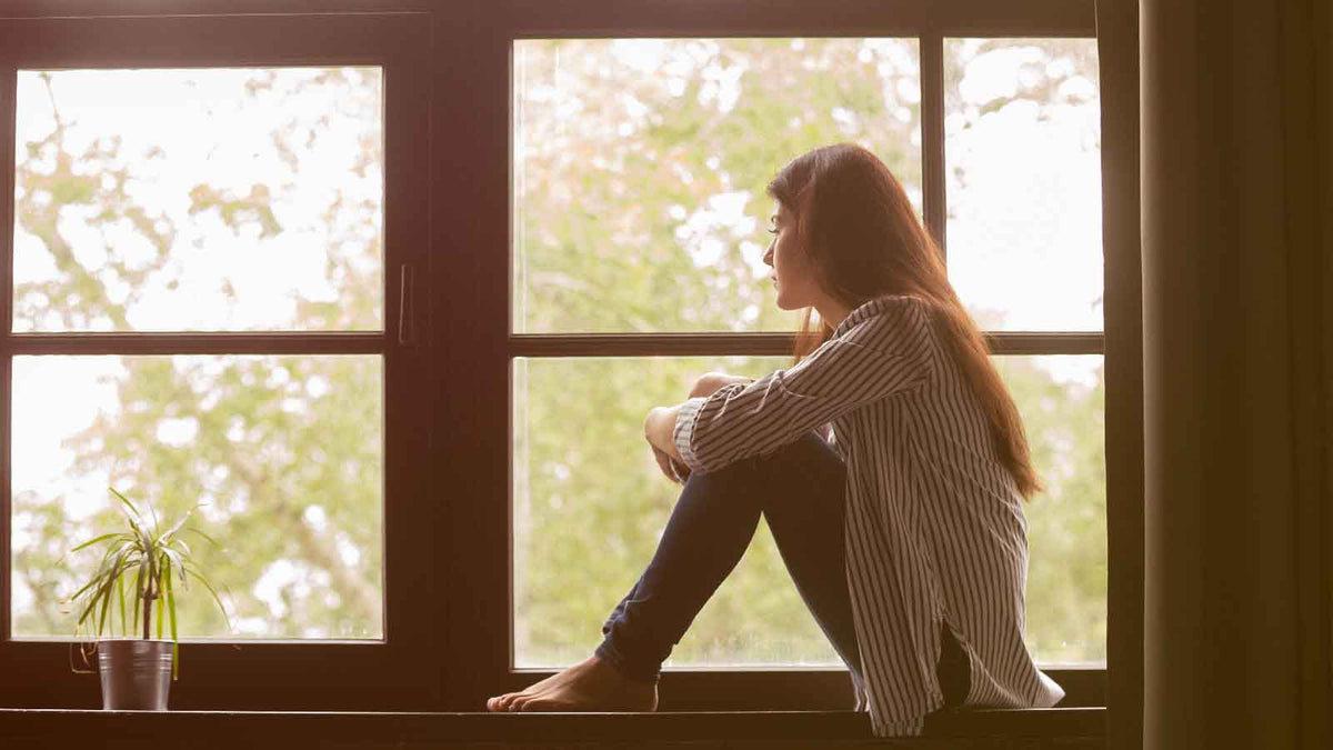 Eine junge Frau sitzt auf einer Fensterbank und schaut traurig aus dem Fenster heraus.
