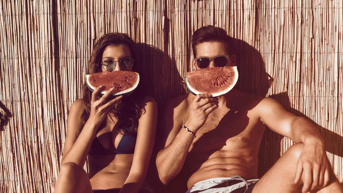 Eine junge Frau und ein junger Mann in Badekleidung halten sich Wassermelonenstücke vor den Mund. Es sieht nach einem Lachen aus.