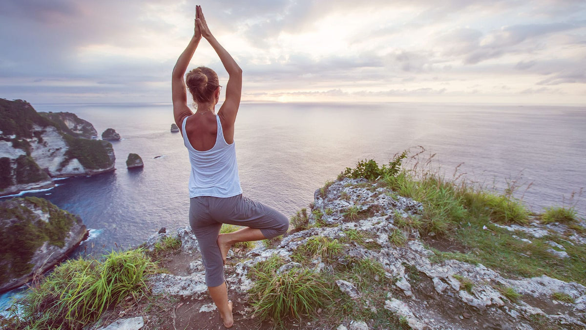 Eine junge Frau befindet sich auf einem Berg am Meer und macht eine Yogaübung - die Baumübung