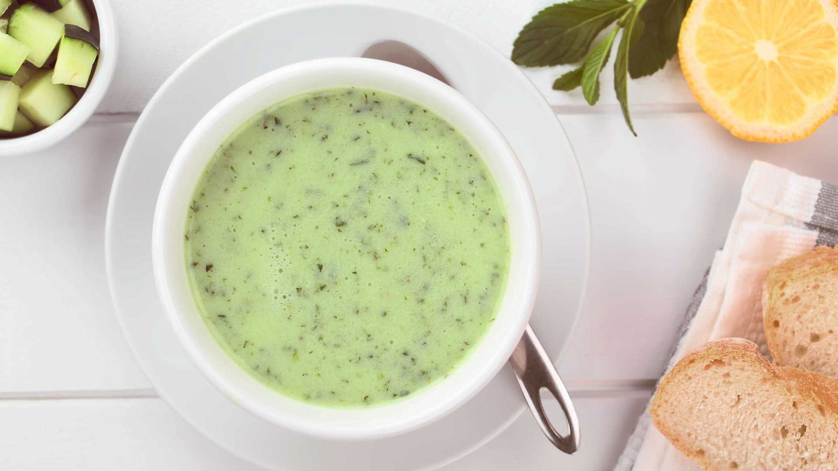 Grüne Gurkensuppe in einer weißen Schale mit Joghurt, Gurkenstückchen, Zitrone und zwei Scheiben Weißbrot