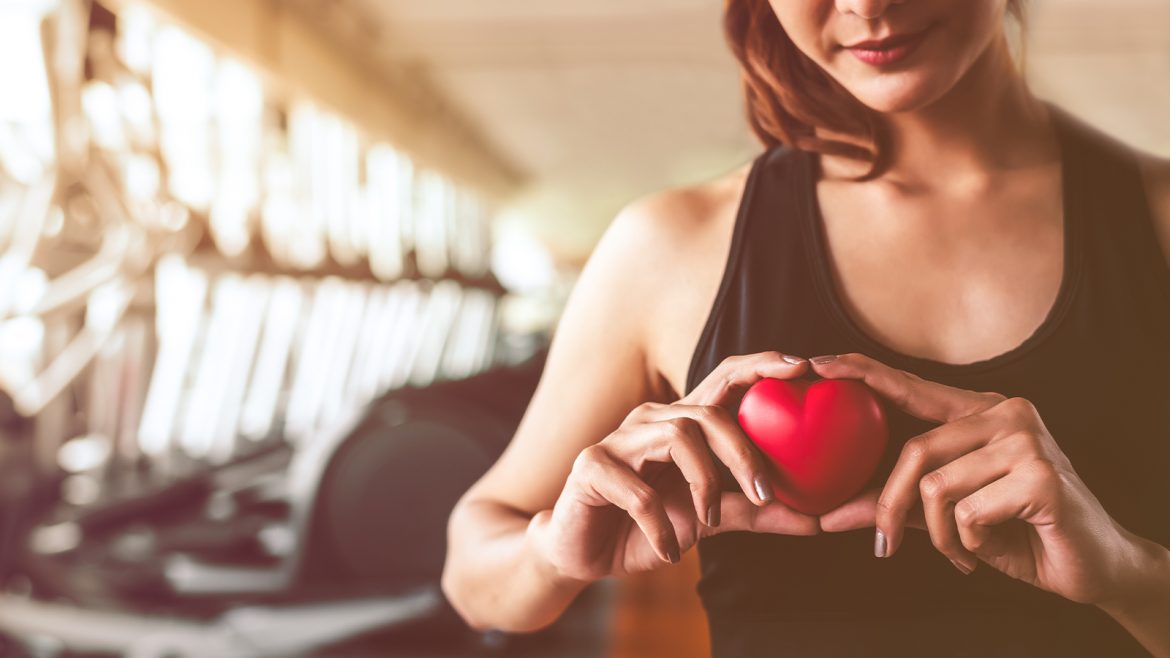Eine Frau hält ein Tomatenherz in der Hand. Im Hintergrund sieht man verschwommen ein Fitnessstudio.