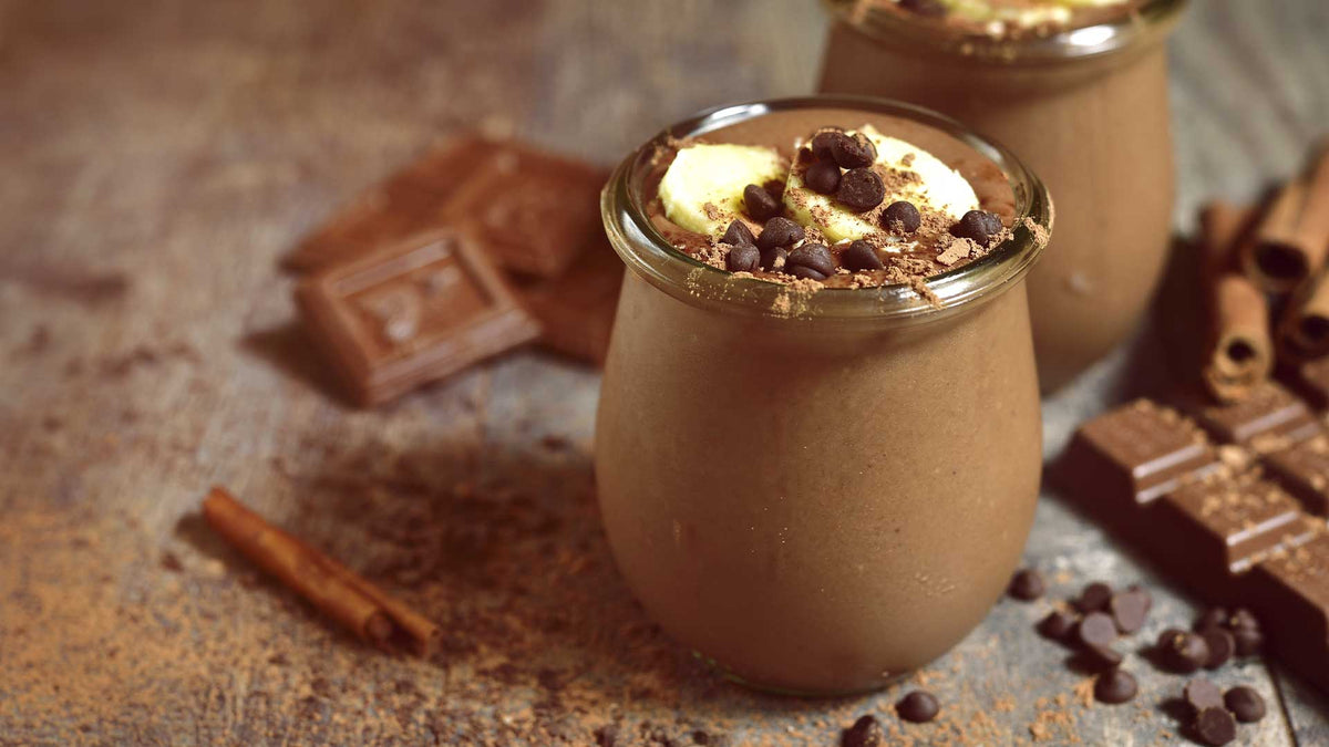 Smoothie-Glas mit Kakannibs und Bananen zubereitet. Zimtstangen, Kakaonibs und Schokoladenriegel als Dekoration um das Smoothieglas gelegt.