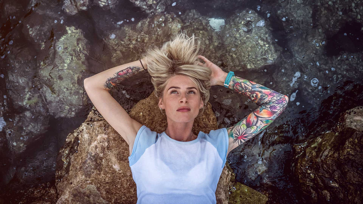 Eine junge tätowierte Frau liegt am Wasser an einem Felsstrand und ist ganz entspannt.