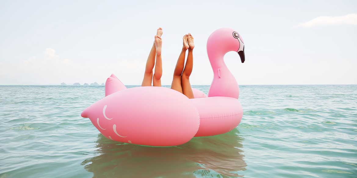 Zwei junge Frauen liegen rücklings in einer Riesen-Flamingo-Luftmatratze im Meer. Man sieht nur ihre nach oben gestreckten Beine.