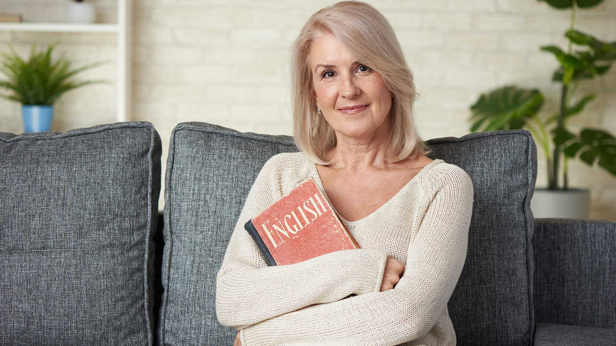 Eine Frau sitzt lächelnd mit einem Englisch-Lernbuch auf dem Sofa