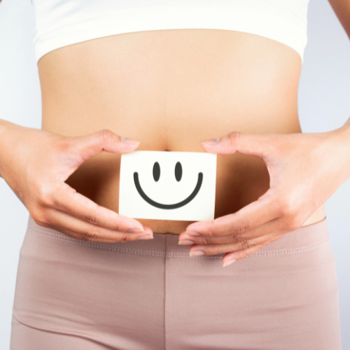 Eine Person hält eine Karte mit einem Smiley-Gesicht über ihren nackten Bauch, das die Gesundheit des Darms symbolisiert.