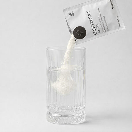 Das geöffnete Sachet von Elektrolyt Aktiv. Das Pulver fällt in ein Glas Wasser