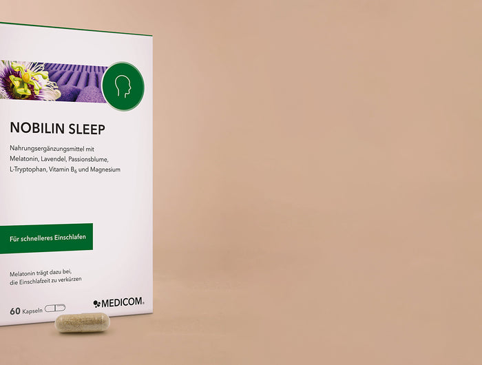 Die Produktverpackung von Nobilin Sleep von Medicom auf der linken Seite im Bild mit einer Kapsel im Vordergrund. Im Hintergrund ist eine Schlafmaske in altrosa.