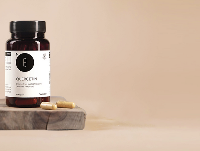 Die Produktdose von Quercetin auf einem Holztablett mit zwei Kapseln im Vordergrund. Ein Glas Wasser hinten links verschwommen.