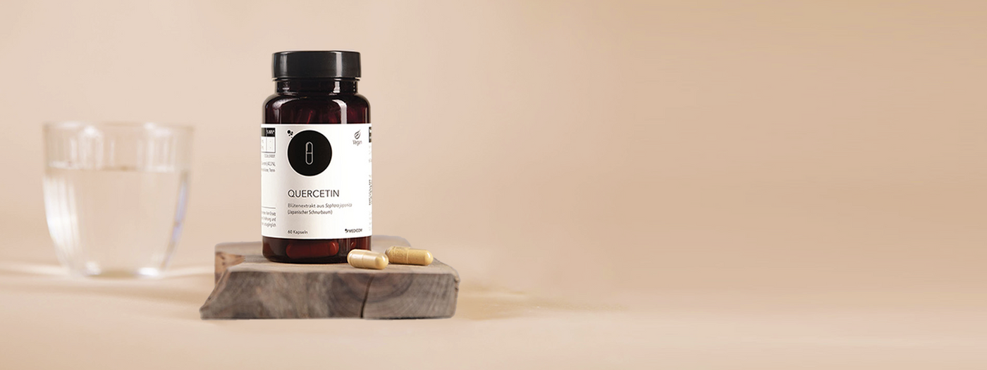 Die Produktdose von Quercetin auf einem Holztablett mit zwei Kapseln im Vordergrund. Ein Glas Wasser hinten links verschwommen.