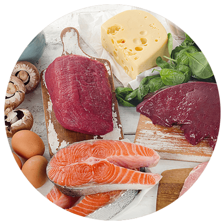 Verschiedene eiweißhaltige Lebensmittel wie rohes Rindfleisch, Fisch, Käse, Eier, Pilze, Spinat und Leber auf einer weißen Oberfläche.
