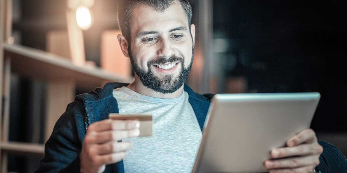 Lächelnder Mann, der eine Kreditkarte in der Hand hält und ein Tablet benutzt, sitzt in einem Raum mit warmer Beleuchtung.