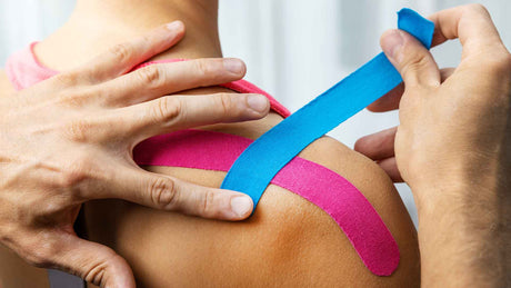 Die Finger eines Physiotherapeuten kleben bunte Tapes auf eine Schulter.