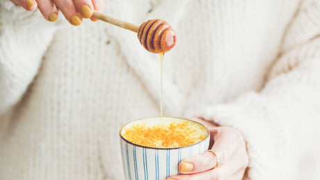 Eine Frau mit einem weißen Pulli hält eine Curcuma Latte mit einem Honiglöffel in der Hand.