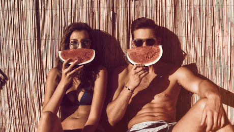 Eine junge Frau und ein junger Mann in Badekleidung halten sich Wassermelonenstücke vor den Mund. Es sieht nach einem Lachen aus.