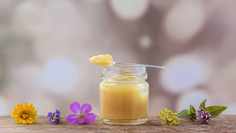 Ein Glas mit Gelée Royale und ein Löffel mit dem königlichen Futtersaft dekoriert mit einer gelben und einer lila Blume.