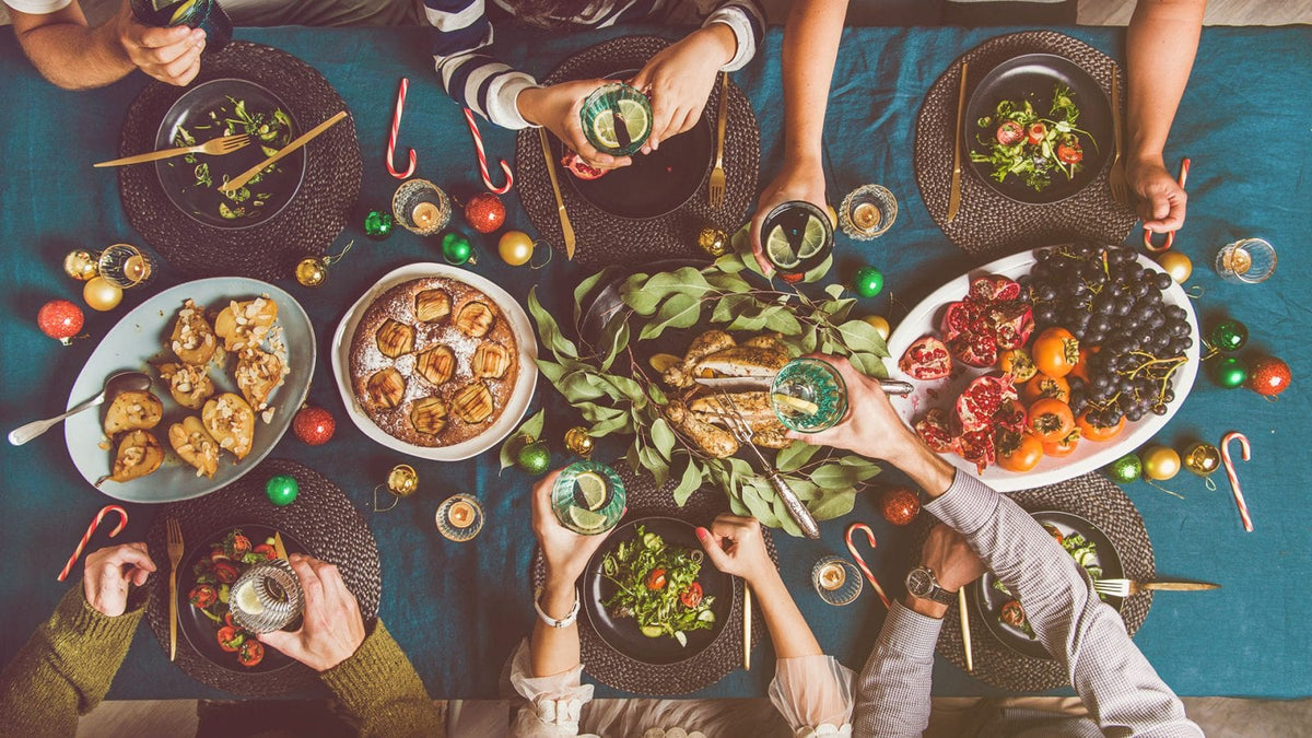 Ein schön dekorierter Weihnachtstisch mit gesunder Kost. Man sieht Arme und Hände der Menschen, die am Tisch sitzen und auf Weihnachten anstoßen.