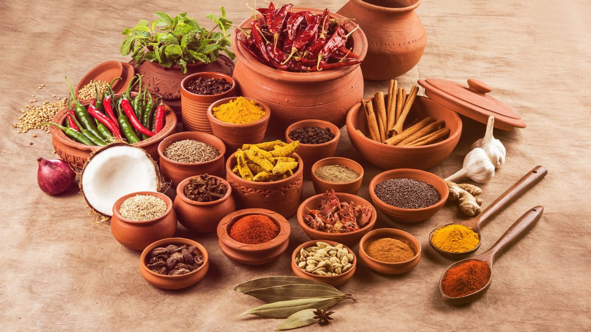 In mehreren Tonschalen befinden sich Gewürzpulver, Kräuter, Chilischoten, Ingwer, Zimtstangen, Knoblauch etc.