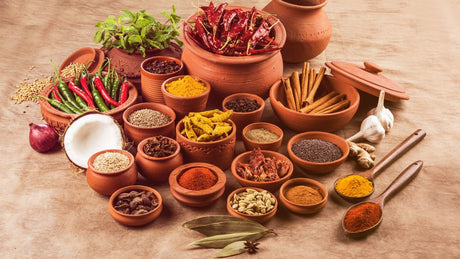 In mehreren Tonschalen befinden sich Gewürzpulver, Kräuter, Chilischoten, Ingwer, Zimtstangen, Knoblauch etc.