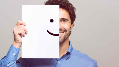 Ein junger Mann lächelt in die Kamera. Er hält sich ein Blatt mit einem halben Smiley auf seine halbe Gesichtshälfte.