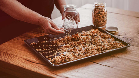 Ein Backblech mit selbstgemachtem Granola. Hände füllen Granola in Glasbehälter.