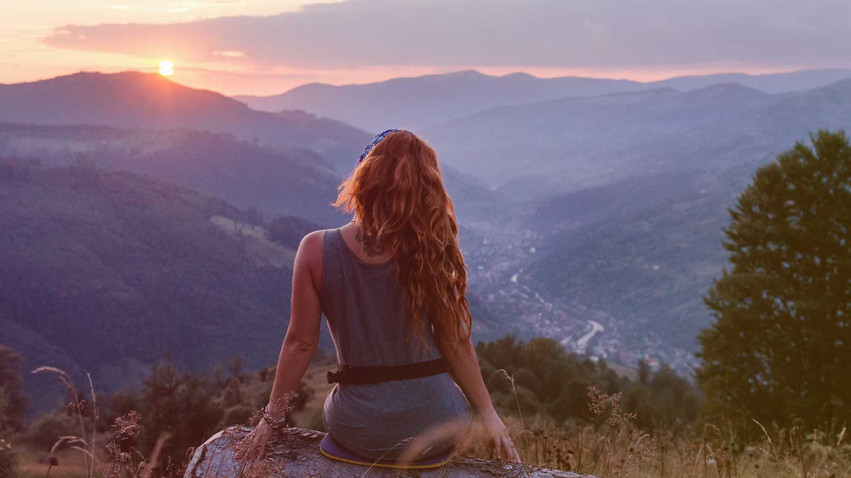 Eine rothaarige Frau sitzt auf einem Berggipfel und schaut auf eine Berglandschaft mit Fluss