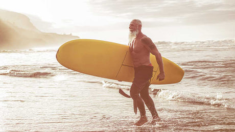 Ein älterer Surfer kommt aus dem Meer. Er hat einen Neoprenanzug halb ausgezogen und hat ein Surfbrett in der Hand