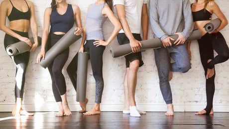 Eine junge Pilatesgruppe steht mit Pilatesmatten in der Hand an der Wand gelehnt.