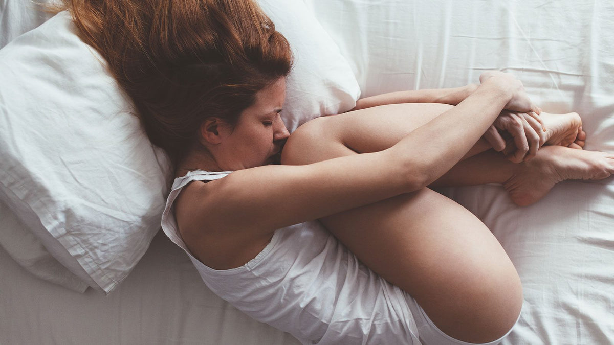 Eien junge Frau in weißem Unterhemd und Unterhose krümmt sich im Bett vor Schmerzen