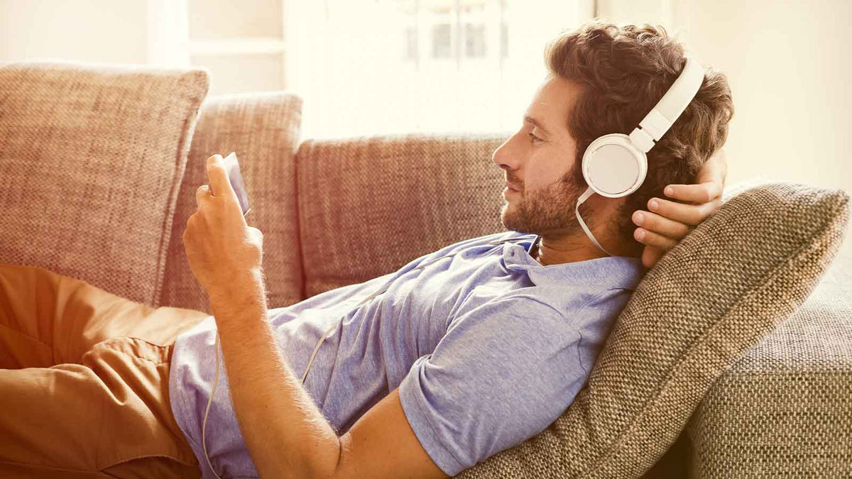 Ein junger Mann sitzt auf dem Sofa, hat Kopfhörer auf und hört einen Podcast.