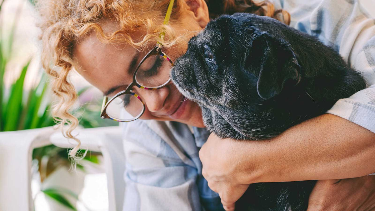 Eine Frau mit Locken und Brille umarmt ihren schwarzen Hund.