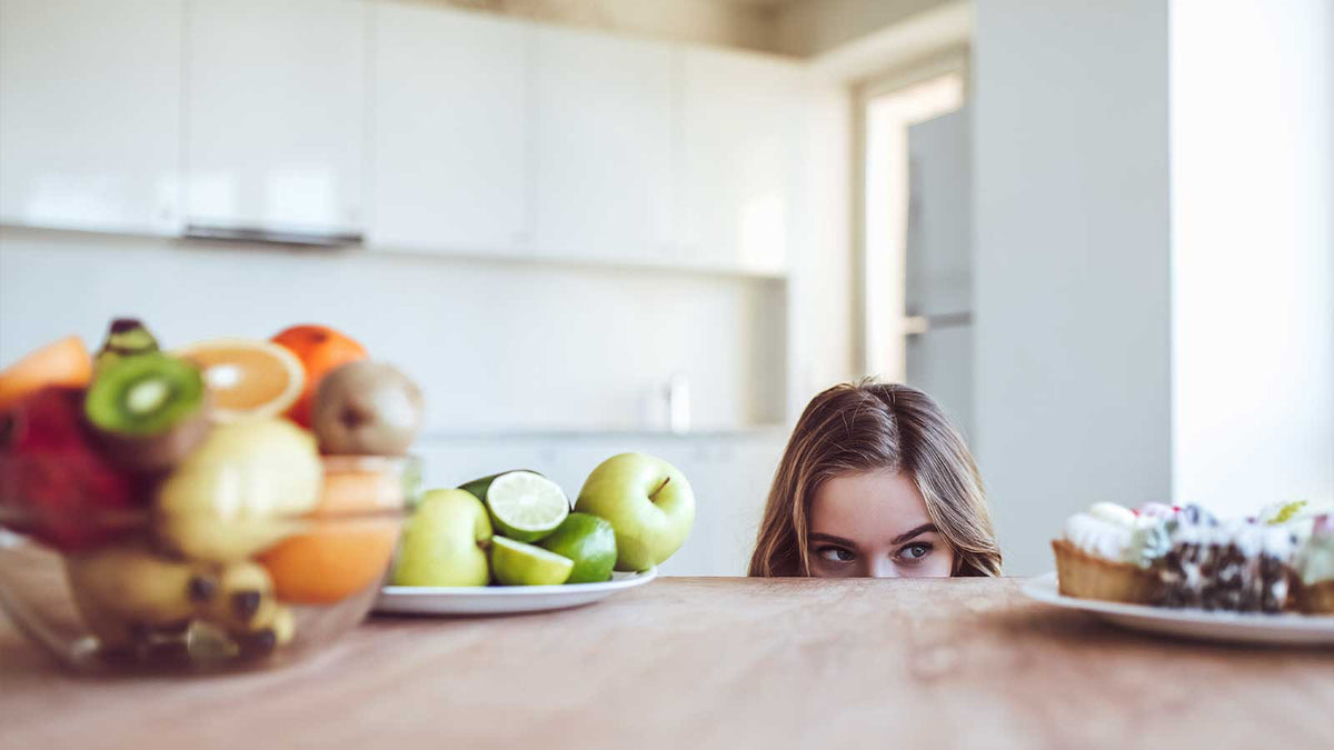 Eine Frau schaut in Augenhöhe auf einen Teller mit Süßigkeiten. Vor ihr liegen auch Obst wie Bananen und Zitrusfrüchte.
