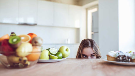 Eine Frau schaut in Augenhöhe auf einen Teller mit Süßigkeiten. Vor ihr liegen auch Obst wie Bananen und Zitrusfrüchte.