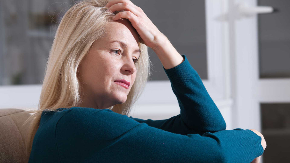 Eine blonde Frau mittleren Alters sitzt erschöpft auf dem Sofa und fasst sich müde mit der Hand an den Kopf