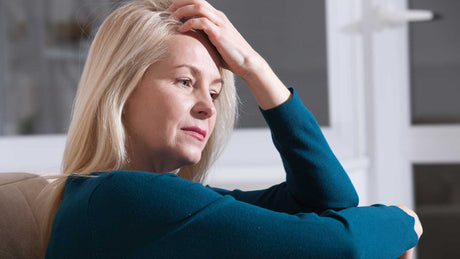 Eine blonde Frau mittleren Alters sitzt erschöpft auf dem Sofa und fasst sich müde mit der Hand an den Kopf