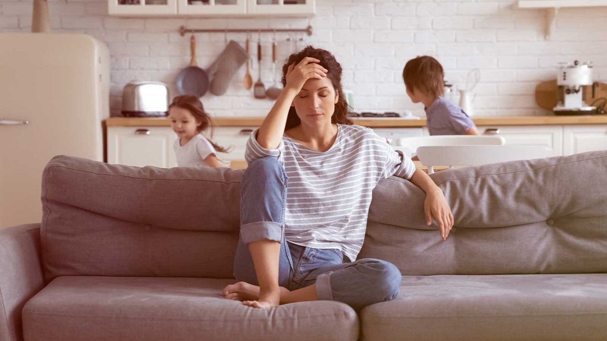 Eine Frau sitzt auf dem Sofa und ist gestresst. Hinter ihr laufen Kinder.