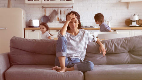 Eine Frau sitzt auf dem Sofa und ist gestresst. Hinter ihr laufen Kinder.