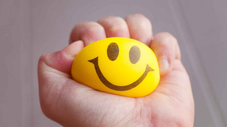 Eine Hand zerdrückt einen gelben kleinen Schaumstoffball mit einem Smiley