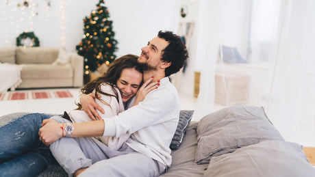 Ein Paar (Frau und Mann) sitzt vor dem Weihnachtsbaum auf dem Sofa und umarmt sich lächelnd.