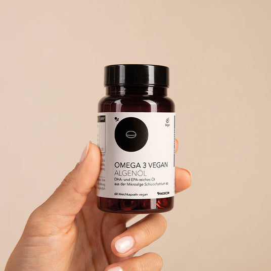 Eine Hand hält eine Flasche mit Omega 3 Vegan Algenöl Softgel-Kapseln vor einem beigen Hintergrund.
