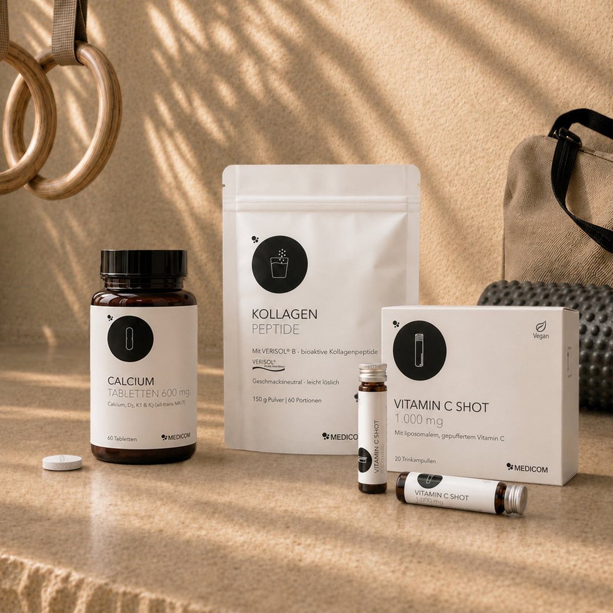 Die Schachteln und Tuben des GLP-1-Begleitsets „Muskeln & Knochen“ von Medicom sind auf einem Tisch ausgestellt, im Hintergrund sind Turnringe zu sehen, die die positive Wirkung des Produkts auf die Gesundheit von Muskeln und Knochen unterstreichen.