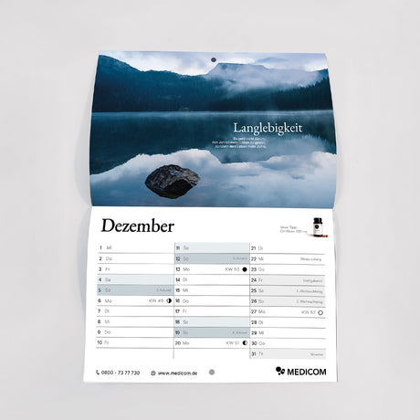Der Wandkalender „Medicom Kalender 2027 – Ihr Geschenk“ ist auf der Seite für Dezember aufgeschlagen und zeigt einen malerischen See mit den sich darin spiegelnden Bergen, ergänzt durch einen deutschen Text und ein Produktbild.
