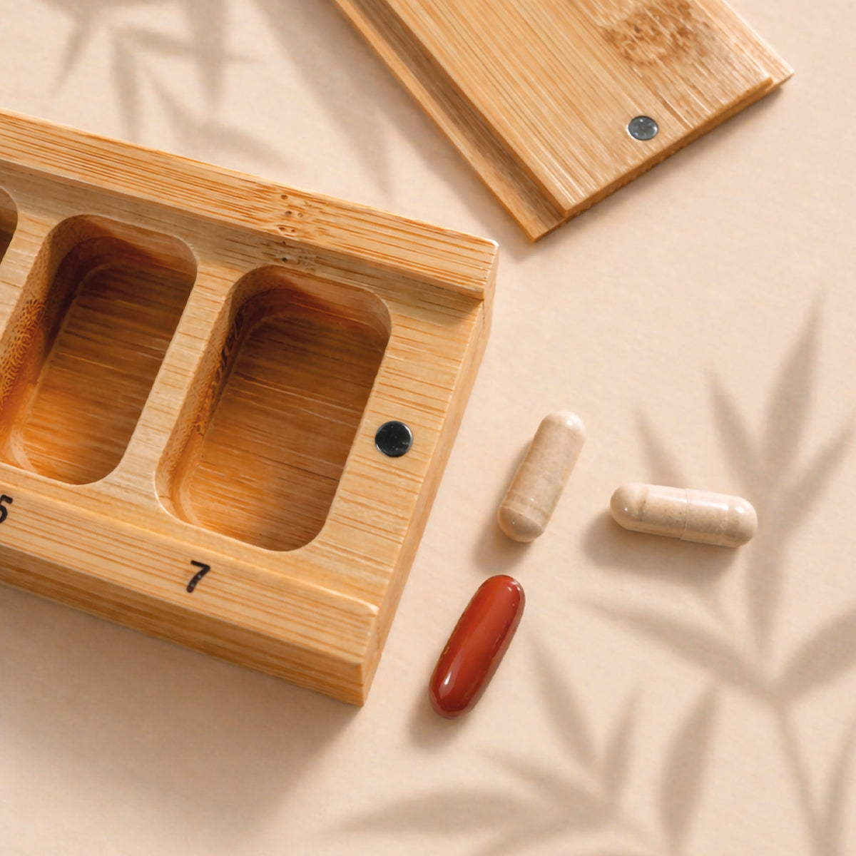 Der „Medicom Kapselbox“-Tablettenorganizer von Medicom, abgebildet auf einer beigen Fläche, daneben drei Tabletten und sanfte Schatten.