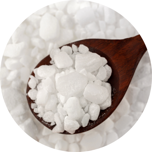 Ein Holzlöffel, der weiße, klobige Kristalle hält, mit weiteren Kristallen im Hintergrund.