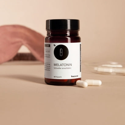 Die Produktdose von Melatonin von Medicom und zwei weiße Kapseln im Vordergrund. Im Hintergrund eine Schlafmaske und eine Glasunterlage mit Kapseln. 