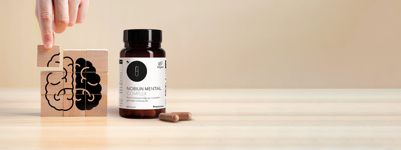 Die Produktdose von Nobilin Mental Complex mit zwei Kapseln vor der Dose. Daneben Holzklötze auf denen ein Gehirn abgebildet ist.