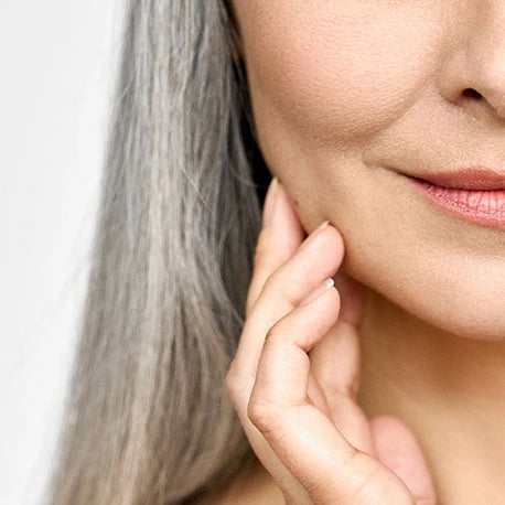 Nahaufnahme einer Frau mit langen grauen Haaren, die ihre Wange berührt und sanft lächelt.