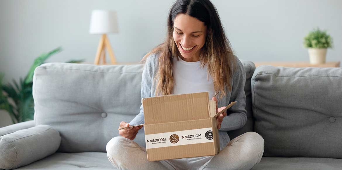 Eine Frau, die auf einer Couch sitzt, lächelt und öffnet einen Karton auf ihrem Schoß.